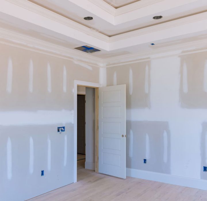 Interior of a room under construction showing unfinished drywall and a sophisticated ceiling design with multiple layers and recessed lighting placements.