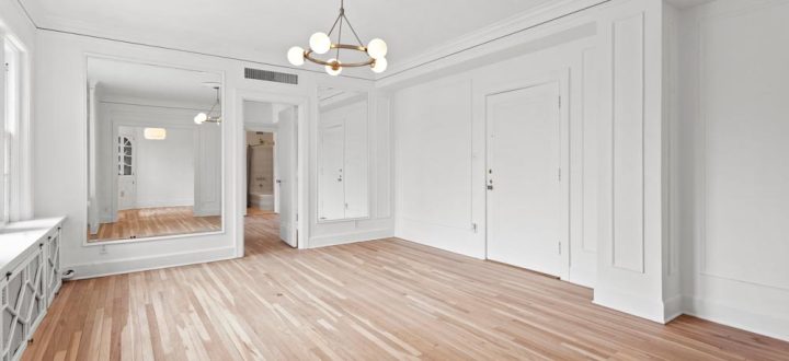 A spacious and bright open-plan living area with polished wooden flooring, white walls, and elegant moldings, featuring a chic chandelier and ample natural light.