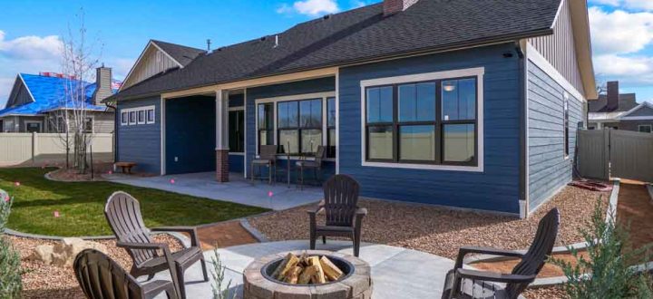 Spacious backyard featuring a blue-gray house with modern landscaping, including a stone patio with wooden Adirondack chairs around a fire pit, perfect for outdoor gatherings.
